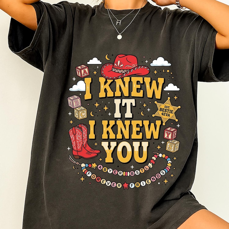 May include: A charcoal gray t-shirt featuring a graphic print with the text I KNEW IT I KNEW YOU. The design includes a red cowboy hat, red cowboy boots, alphabet blocks, a sheriff star badge labeled EST. BESTIE 4EVA, and a beaded necklace graphic.