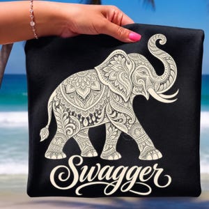 May include: A black t-shirt featuring a large, off-white graphic of an elephant decorated with intricate floral and paisley patterns. Below the elephant, the word "Swagger" is printed in a large, elegant, white cursive script font.