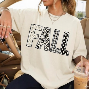 May include: A cream-colored short-sleeve t-shirt featuring the word FALL printed in large block letters. Each letter has a distinct black and white pattern: floral, daisy, buffalo plaid, and polka dot designs fill the letters.