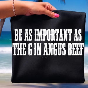 May include: A black t-shirt featuring the white text BE AS IMPORTANT AS THE G IN ANGUS BEEF printed in a bold, serif font across the front. The garment is made of a solid black fabric and displays the text clearly in two lines of capitalized letters.