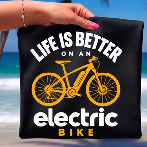 Puede incluir: Una camiseta negra con un gráfico amarillo de una bicicleta eléctrica centrado entre el texto blanco "LIFE IS BETTER ON AN" en la parte superior y "electric BIKE" en la parte inferior. El diseño está impreso en una fuente negrita y clara.