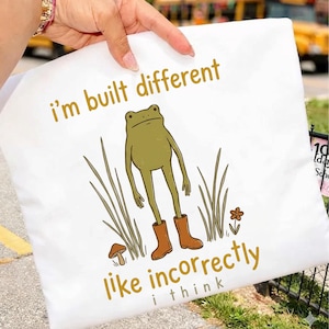 May include: A white t-shirt featuring a graphic of a green frog wearing brown boots, standing among tall grass and a small mushroom. The text reads i'm built different above the frog and like incorrectly i think below the frog in gold lettering.
