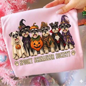 Puede incluir: Una camiseta rosa claro con un gráfico de seis perros Schnauzer vestidos con disfraces de Halloween, incluyendo un fantasma, una calabaza, un espantapájaros, un vampiro y una bruja. El texto debajo dice SPOOKY SCHNAUZER SOCIETY.