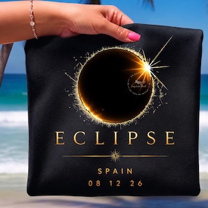 Può includere: Un articolo in tessuto nero con una grafica dorata di un'eclissi solare e un effetto a stella brillante. Il testo recita ECLIPSE, SPAIN e 08 12 26 in caratteri dorati. Una sottile linea orizzontale con un'icona a stella centrale separa il testo.