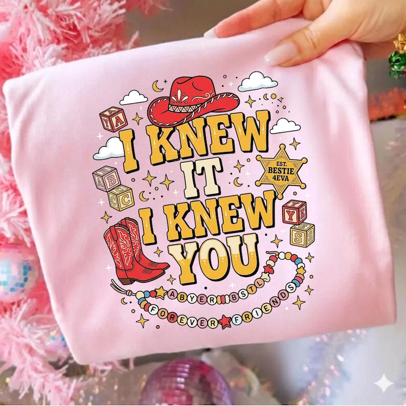 May include: A light pink t-shirt featuring a graphic print with the text I KNEW IT I KNEW YOU in yellow. The design includes a red cowboy hat, red cowboy boots, alphabet blocks, a sheriff star badge, and a colorful beaded necklace with text.