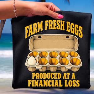 Könnte beinhalten: Ein schwarzes T-Shirt mit einer Grafik eines offenen Eierkartons, der ein einzelnes weißes Ei enthält. Der Text darüber lautet FARM FRESH EGGS in Gelb, und der Text darunter lautet PRODUCED AT A FINANCIAL LOSS in Gelb auf dunklem Hintergrund.