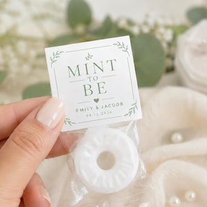 May include: A white ring-shaped mint candy in a clear plastic wrapper with a square paper topper. The topper features green text that reads MINT TO BE, EMILY & JACOOB, and the date 09.12.2026, framed by delicate green leaf illustrations.