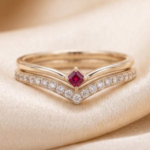 May include: A gold ring set consisting of two bands. The top band features a princess-cut red ruby at the center. The bottom band is a chevron-shaped eternity style ring set with a row of small, clear, round-cut white stones along the V-shape.