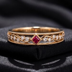 May include: A gold band ring featuring a central princess-cut red ruby flanked by small round white stones set within a delicate swirling metal design. The edges of the ring are detailed with a fine beaded texture, creating a refined appearance.