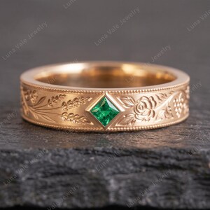 May include: A rose gold band ring featuring a central princess-cut green gemstone set in a diamond-shaped bezel. The band is decorated with engraved floral and wheat motifs, framed by delicate beaded edges along the top and bottom borders.