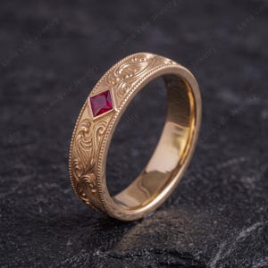 May include: A rose gold band ring featuring an intricate engraved scrollwork pattern along the outer surface and a single square-cut red gemstone set in a diamond-shaped frame. The metal has a polished finish with beaded edges.