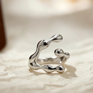 Peut inclure: Une bague en argent poli présentant un design ouvert, irrégulier et fluide. L'anneau se compose de segments lisses, arrondis et aux formes organiques qui créent une silhouette continue, ondulée et asymétrique avec une finition métallique.