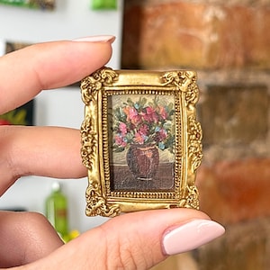 May include: A miniature rectangular painting in a decorative gold-colored frame. The artwork depicts a dark brown vase filled with a vibrant bouquet of pink, red, and green flowers, set against a neutral background. The frame features ornate, raised detailing.