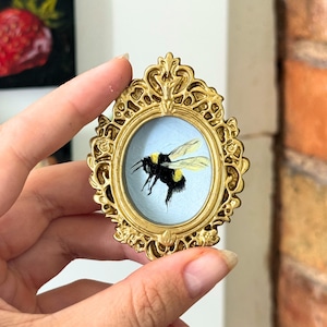 Puede incluir: Un pequeño marco ovalado de color dorado con intrincados detalles decorativos de volutas. Dentro del marco hay una pintura en miniatura sobre un fondo azul claro que muestra un abejorro negro y amarillo con alas translúcidas en vuelo.