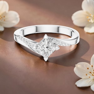 May include: A silver-coloured ring featuring a central princess-cut clear gemstone held by four prongs. The band is accented with a row of small, round, clear stones embedded along each side of the shank, leading up to the central setting.