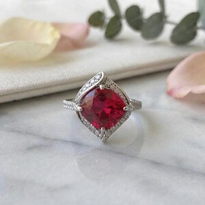May include: A silver-toned ring featuring a large, cushion-cut, vibrant red gemstone at its center. The stone is surrounded by a halo of small, clear, sparkling stones set in a diamond-shaped frame, with additional small stones along the band.