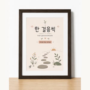 May include: A framed wall art print featuring a beige background with a path of stepping stones leading toward a rising sun. The text reads "한 걸음씩", "han georeumssik", and "step by step" in black and brown, accompanied by small botanical illustrations.