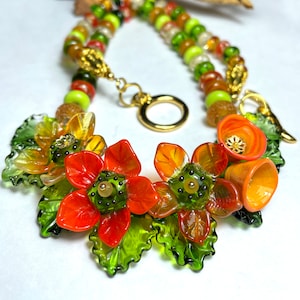 Può includere: Una collana con una catena d'oro e perline di vetro verde e arancio. La collana presenta un gruppo di fiori di vetro arancio e verde.