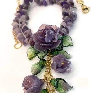May include: A necklace featuring a strand of tumbled purple amethyst beads with a gold-tone chain. The focal point is a cluster of translucent purple glass flowers and green glass leaves attached to the gold chain, finished with a lobster clasp.