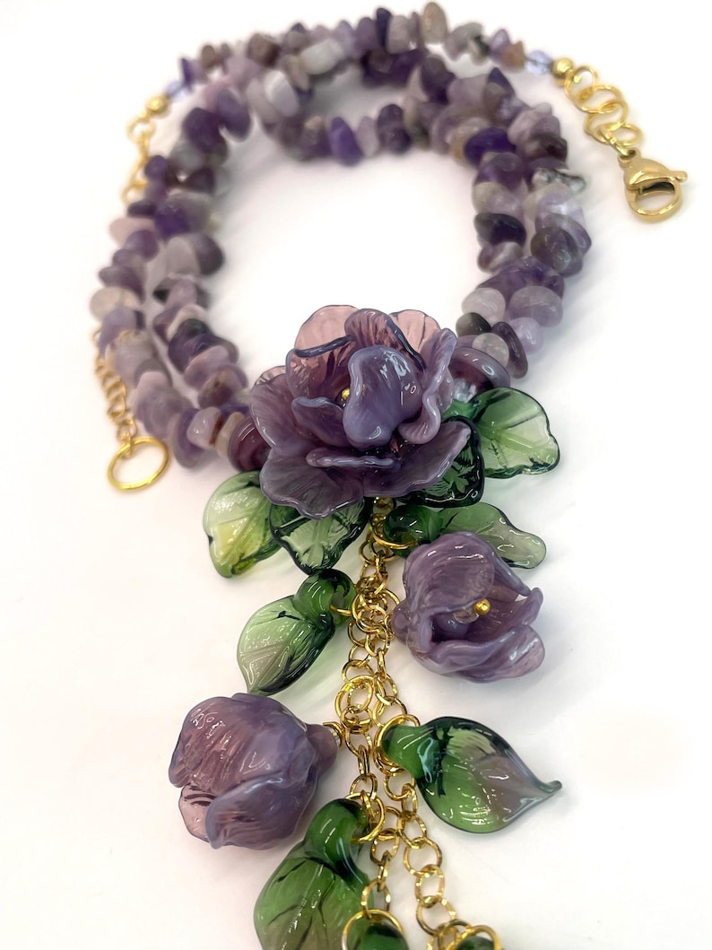 May include: A necklace featuring a strand of tumbled purple amethyst beads with a gold-tone chain. The focal point is a cluster of translucent purple glass flowers and green glass leaves attached to the gold chain, finished with a lobster clasp.