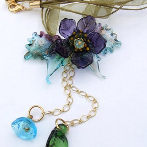 Può includere: Una delicata collana a catena dorata con un ciondolo floreale. Il ciondolo presenta un fiore viola con un centro verde e foglie blu e verdi. La catena è attaccata a un nastro marrone.