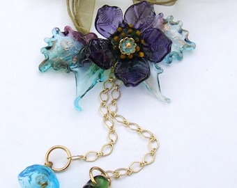 May include: A delicate gold chain necklace with a floral pendant. The pendant features a purple flower with a green center and blue and green leaves. The chain is attached to a brown ribbon.