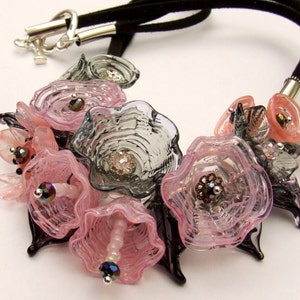 Può includere: Una collana di fiori di vetro rosa e grigio con un cordoncino in pelle scamosciata nero. La collana presenta più fiori di vetro con dettagli intricati e accenti iridescenti.