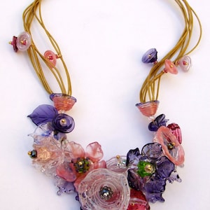 Può includere: Una collana multifilo in oro con perline a forma di fiore in vetro rosa, viola e trasparente. La collana presenta una varietà di forme e dimensioni di fiori, con alcune perline che hanno un effetto a strati.