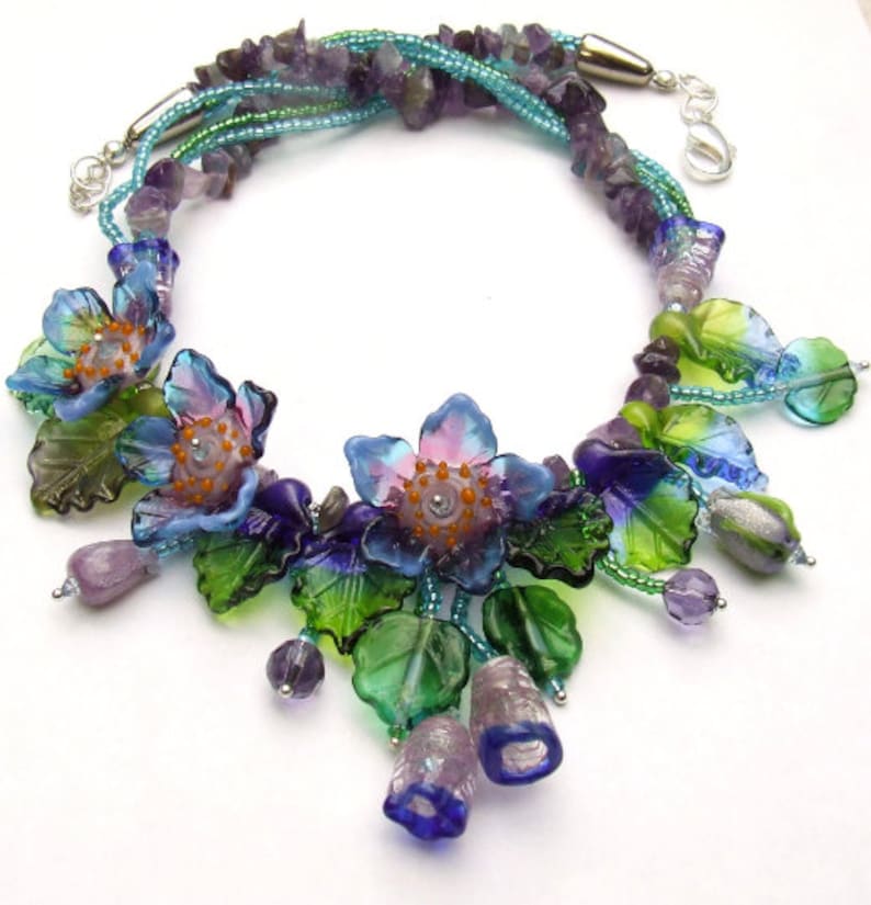 Pu&ograve; includere: Una collana multifilo con perline di ametista e fiori di vetro. I fiori sono blu, verdi e viola con un centro rosa. La collana &egrave; impreziosita da perline argentate e una chiusura argentata.