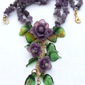 May include: A necklace featuring strands of purple amethyst chip beads and a central pendant arrangement of three carved purple glass roses and several green glass leaves, all connected by gold-toned metal chains and a lobster claw clasp.