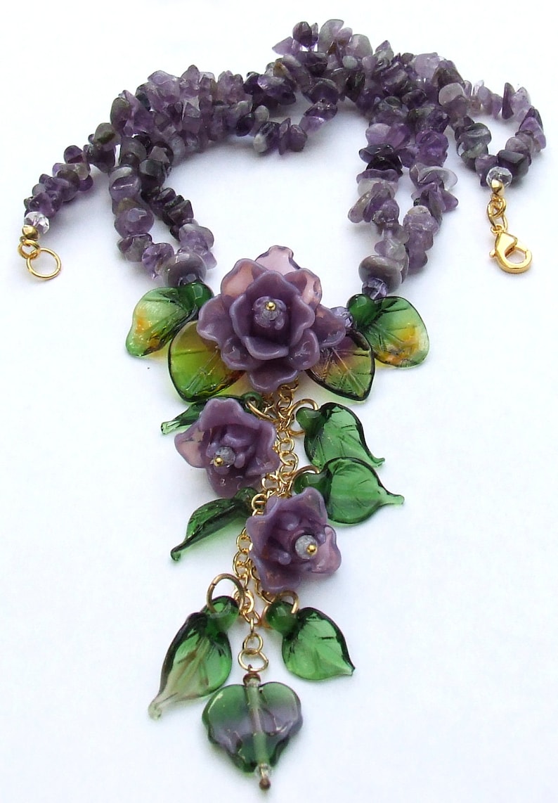 May include: A necklace featuring strands of purple amethyst chip beads and a central pendant arrangement of three carved purple glass roses and several green glass leaves, all connected by gold-toned metal chains and a lobster claw clasp.