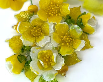 May include: A collection of yellow and white glass flowers with green leaves. The flowers have detailed petals and a textured centre. Additional yellow pansies are in the background. The glass flowers are likely a brooch or decorative item.