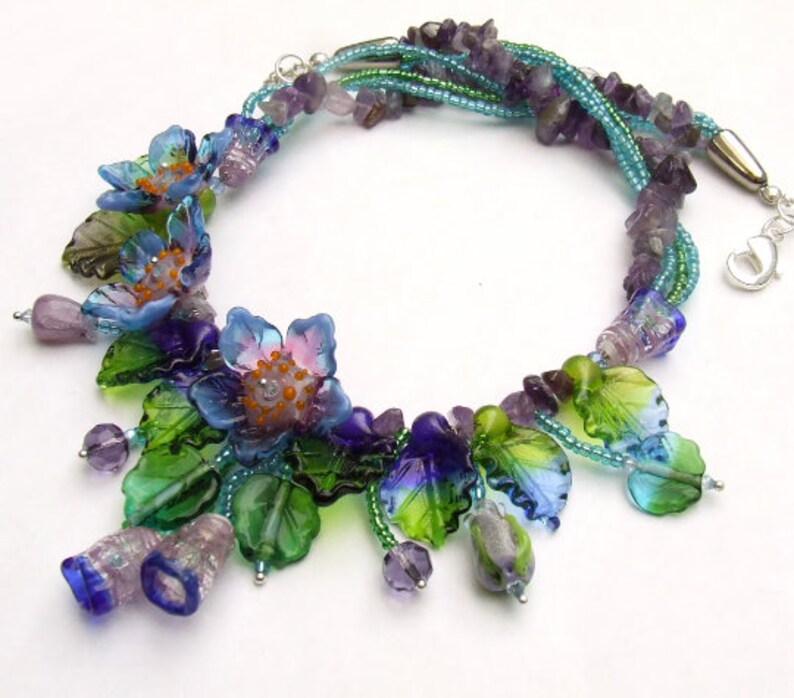 Pu&ograve; includere: Una collana multifilo con perline di ametista e fiori e foglie di vetro in tonalit&agrave; di blu, verde e viola.