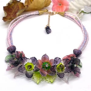 Può includere: Una collana delicata con un designo floreale. La collana è realizzata con perline di vetro rosa, viola e verde. I fiori sono disposti a grappolo e hanno un aspetto vintage. La collana è perfetta per aggiungere un tocco di eleganza a qualsiasi outfit.