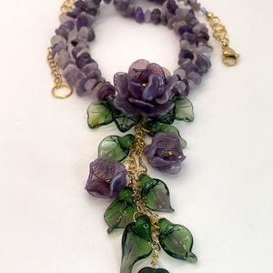 May include: A necklace featuring a strand of tumbled purple amethyst chips and a gold-tone metal chain. The centerpiece consists of a cluster of purple glass flowers and translucent green glass leaves hanging from gold-tone metal links.