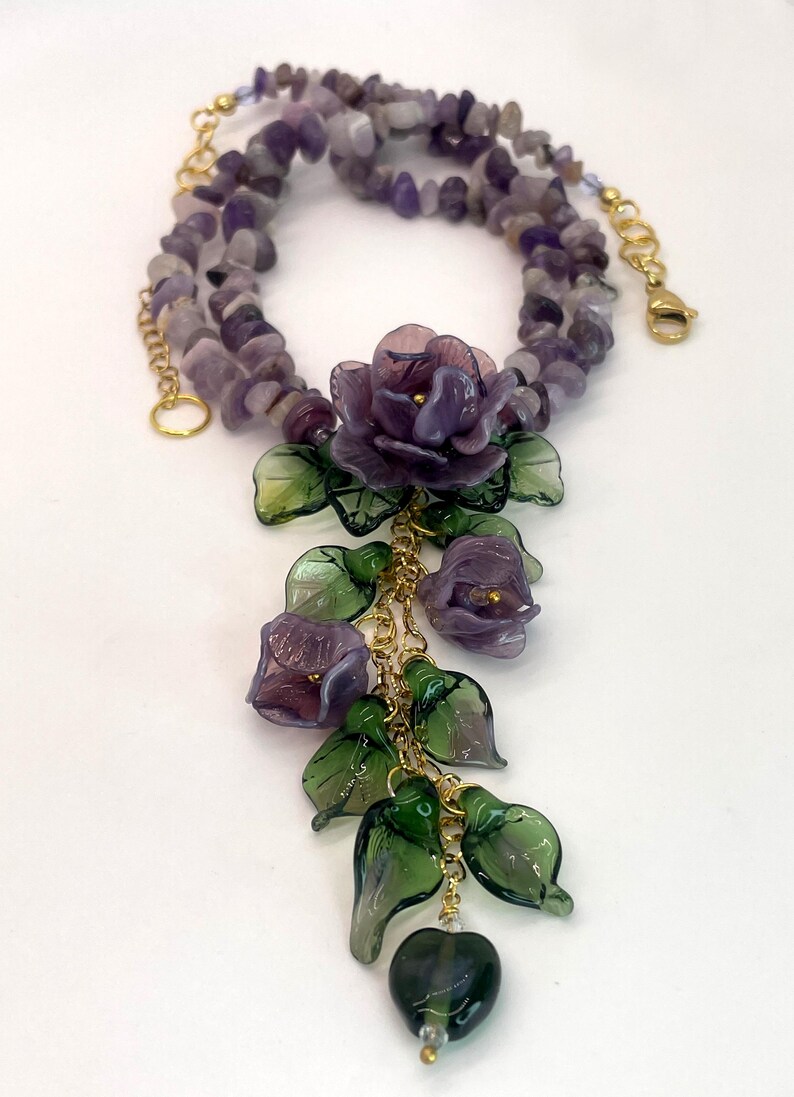 May include: A necklace featuring a strand of tumbled purple amethyst chips and a gold-tone metal chain. The centerpiece consists of a cluster of purple glass flowers and translucent green glass leaves hanging from gold-tone metal links.