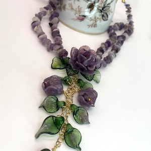 May include: A multi-strand necklace featuring purple amethyst chip beads and a central pendant arrangement of carved purple glass flowers and green glass leaves on gold-tone metal chains, with a dark green heart-shaped bead at the bottom.