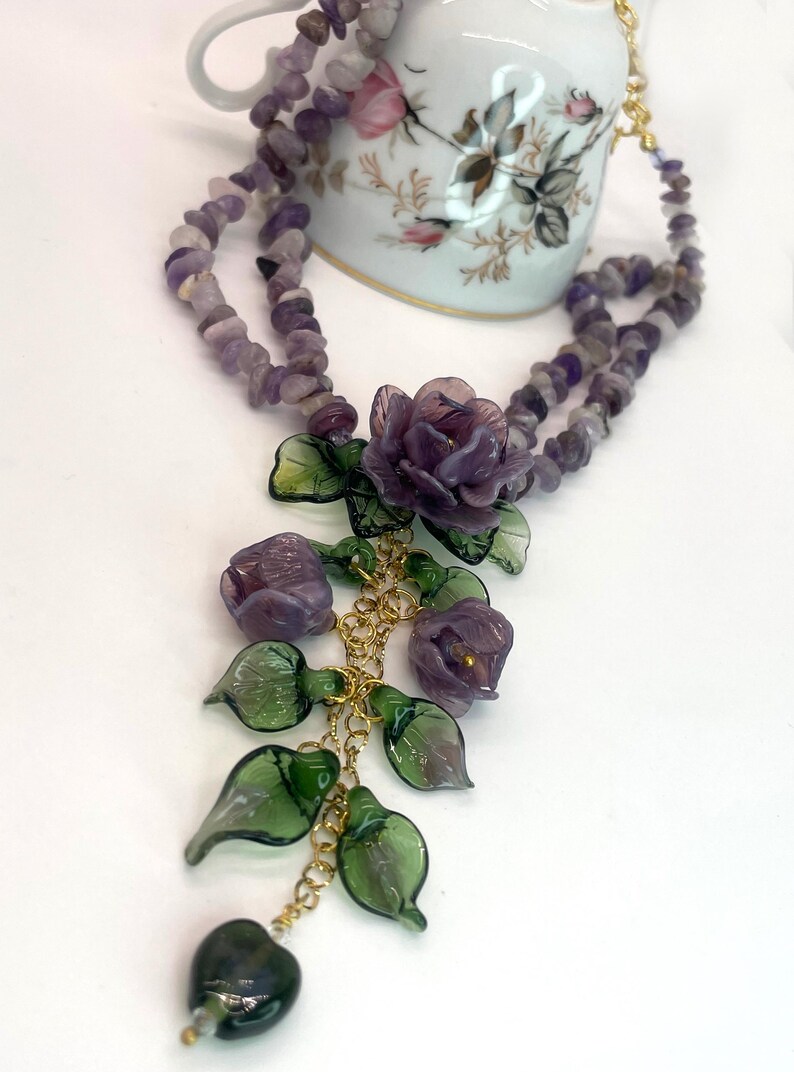 May include: A multi-strand necklace featuring purple amethyst chip beads and a central pendant arrangement of carved purple glass flowers and green glass leaves on gold-tone metal chains, with a dark green heart-shaped bead at the bottom.