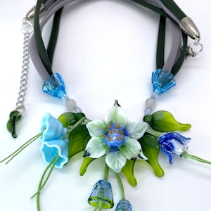 Può includere: Una collana delicata con un gruppo di fiori di vetro blu e bianco con foglie verdi. La collana è realizzata con un cordoncino in tessuto grigio e verde.