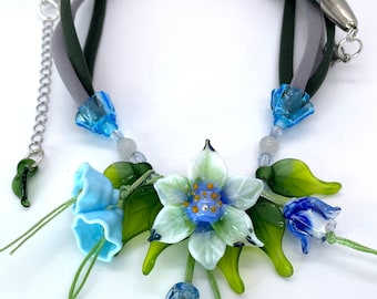 May include: A delicate necklace featuring a cluster of blue and white glass flowers with green leaves. The necklace is made with a gray and green fabric cord.