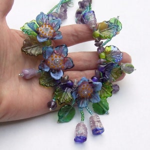 Pu&ograve; includere: Una collana delicata con fiori di vetro in tonalit&agrave; di blu, viola e verde. I fiori sono impreziositi da piccole foglie e perline di vetro. La collana &egrave; infilata su una catena d'argento.