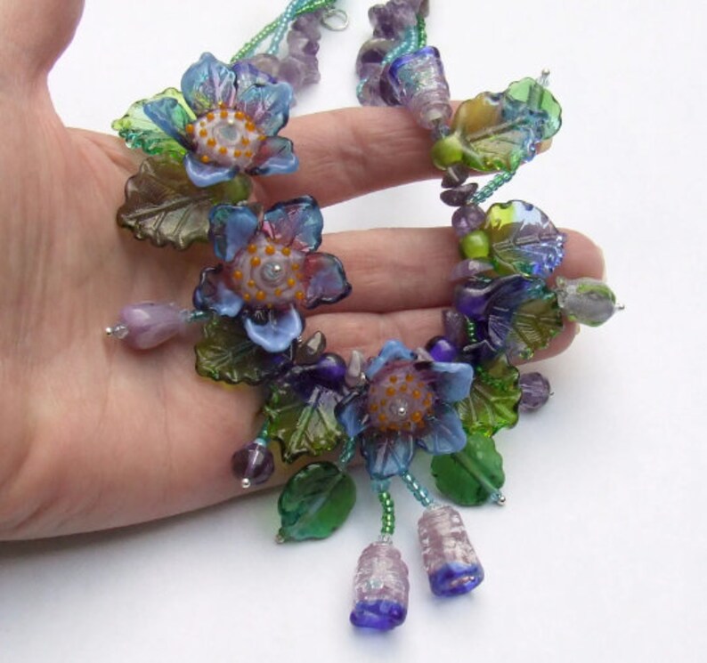 Pu&ograve; includere: Una collana delicata con fiori di vetro in tonalit&agrave; di blu, viola e verde. I fiori sono impreziositi da piccole foglie e perline di vetro. La collana &egrave; infilata su una catena d'argento.