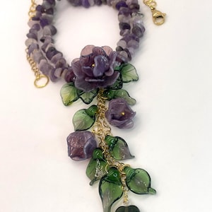 May include: A necklace featuring a strand of tumbled purple amethyst chips and a gold-tone chain. The pendant consists of three translucent purple glass flowers and several green glass leaves attached to gold-tone metal links and a lobster clasp.