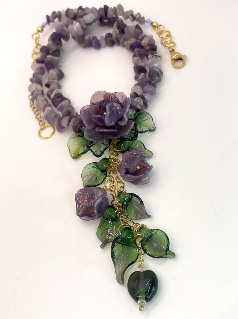 May include: A necklace featuring a strand of tumbled purple amethyst chips and a gold-tone chain. The pendant consists of three translucent purple glass flowers and several green glass leaves attached to gold-tone metal links and a lobster clasp.
