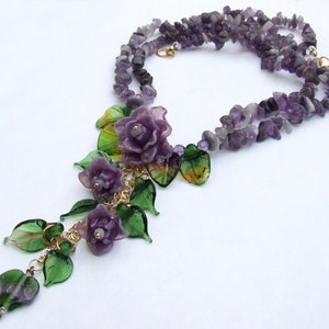 May include: A necklace featuring strands of purple amethyst chip beads. The focal point is a cluster of three purple glass roses and several translucent green glass leaves attached to a gold-tone metal chain, creating a floral pendant design.