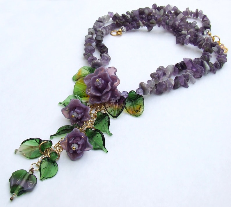May include: A necklace featuring strands of purple amethyst chip beads. The focal point is a cluster of three purple glass roses and several translucent green glass leaves attached to a gold-tone metal chain, creating a floral pendant design.