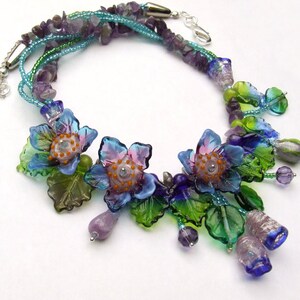 Pu&ograve; includere: Una collana multifilo con perline di vetro blu, verde e viola, tra cui perline a forma di fiore con centri arancioni. La collana &egrave; realizzata con una variet&agrave; di perline, tra cui piccole perline rotonde, perline pi&ugrave; grandi a forma di fiore e perline a forma di foglia.