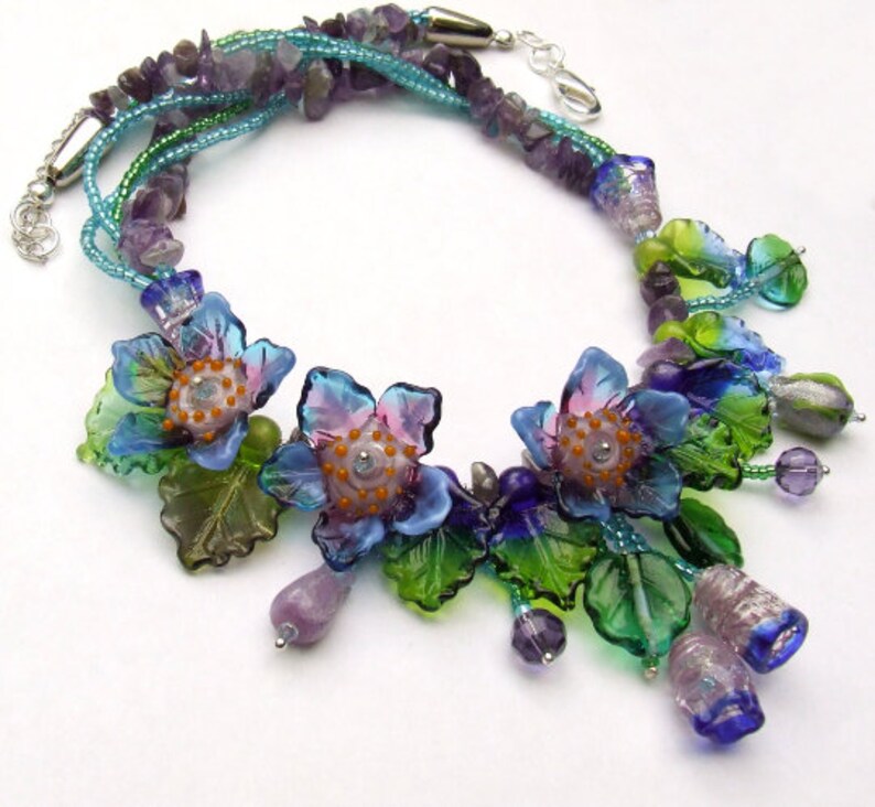 Pu&ograve; includere: Una collana multifilo con perline di vetro blu, verde e viola, tra cui perline a forma di fiore con centri arancioni. La collana &egrave; realizzata con una variet&agrave; di perline, tra cui piccole perline rotonde, perline pi&ugrave; grandi a forma di fiore e perline a forma di foglia.