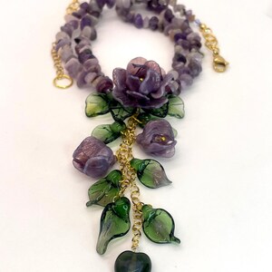May include: A necklace featuring a strand of purple amethyst chip beads and a gold-tone chain pendant. The pendant displays three purple glass flowers, several translucent green glass leaves, and a single dark green heart-shaped glass bead.