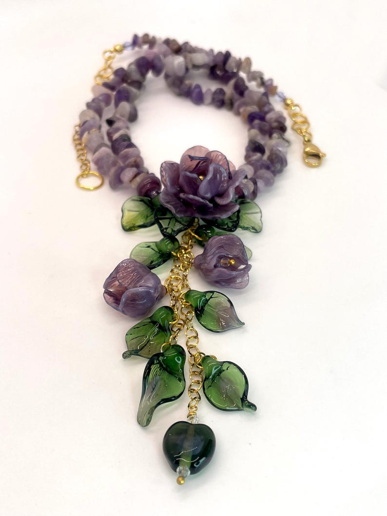 May include: A necklace featuring a strand of purple amethyst chip beads and a gold-tone chain pendant. The pendant displays three purple glass flowers, several translucent green glass leaves, and a single dark green heart-shaped glass bead.